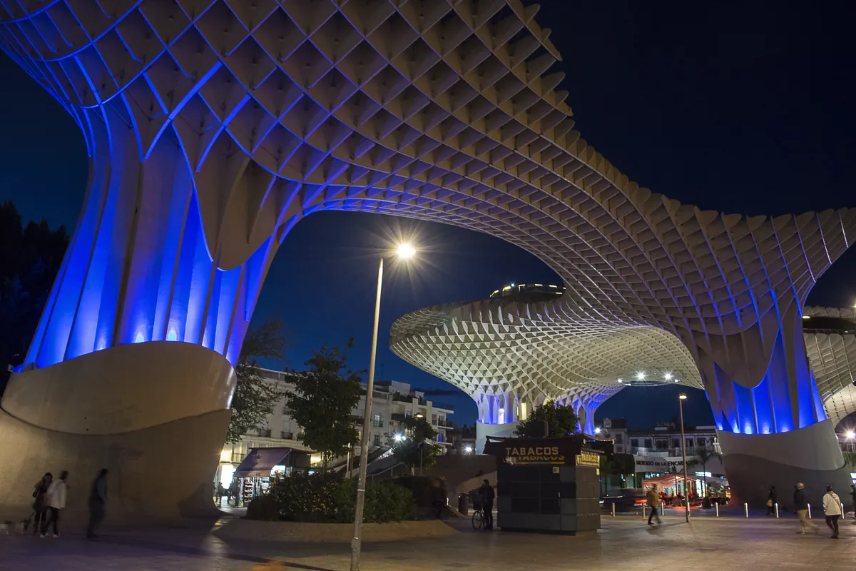 Two of the Metropol Parasol's timber columns lit in blue above a pedestrian street at night in Seville, with the lattice roof overhead