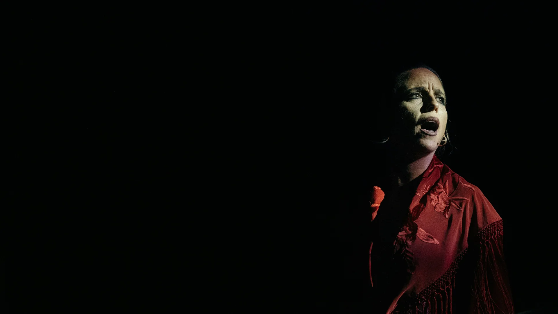 Flamenková zpěvačka v červeném vyšívaném šátku, osvětlená jediným reflektorem na černém jevišti, uprostřed písně s otevřenými ústy